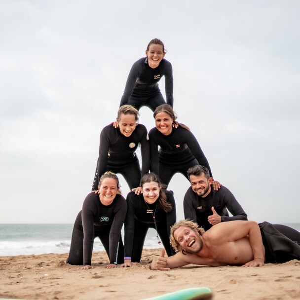 Surf-Schüler bilden eine Menschenpyramide am Strand, Coach zeigt Daumen hoch