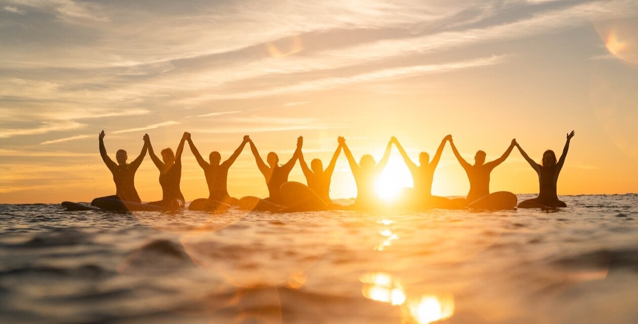 Personen sitzen bei Sonnenuntergang auf Surfbrettern und halten sich an den Händen
