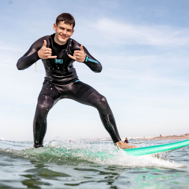 Jugendlicher Surfer im Neoprenanzug zeigt Daumen hoch zur Kamera