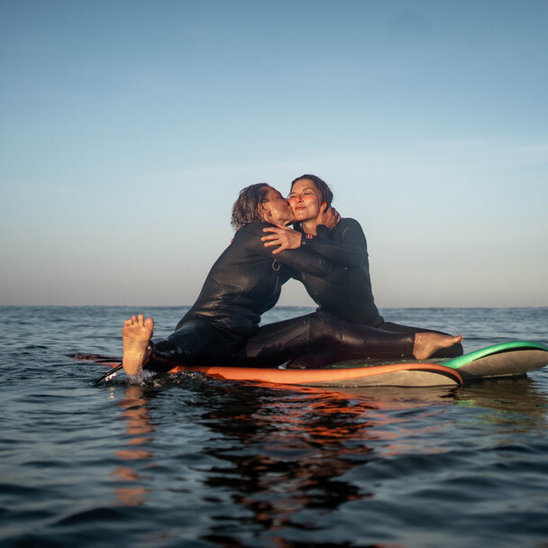 Zwei Mädchen sitzen auf Surfboards, eine gibt der anderen einen spielerischen Kuss