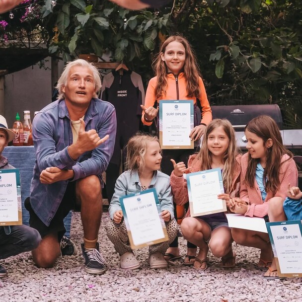 Arne kniet im Innenhof von A-Frame mit Kindern, die Surf-Zertifikate halten