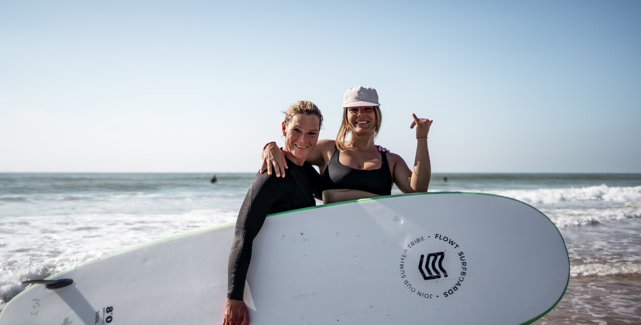 Zwei Frauen mit Surfbrett machen Hangloose Zeichen, Surf & Yoga Retreats