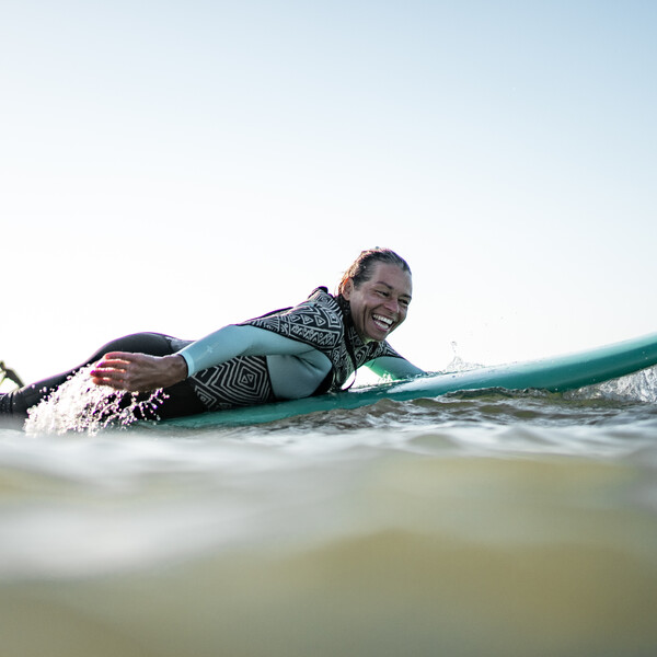 Frau liegt lachend auf Surfbrett und paddelt, Surfcamps für Erwachsene