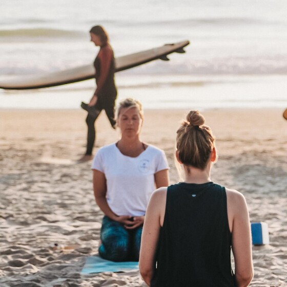 Yoga am Strand, Surferin im Hintergrund, Surfcamp für Erwachsene