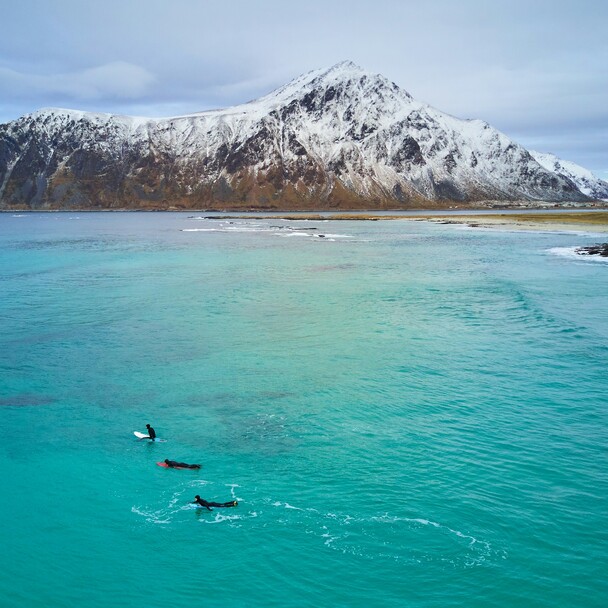 Surfing in Norway, surfers in turquoise water, mountains with snow in the background