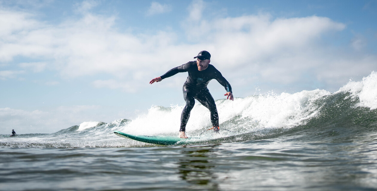 Männlicher Surfer mit Hut reitet eine Welle auf einem Soft-Top-Surfbrett