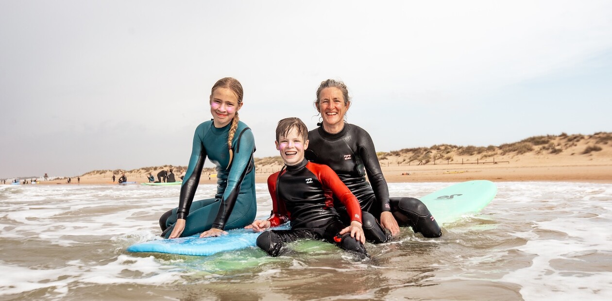 Mutter und zwei Kinder sitzen auf Surfbrettern und lächeln in die Kamera