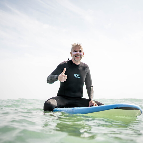 Learning to surf at 50, older man sits on surf bed in the water and points thumbs up