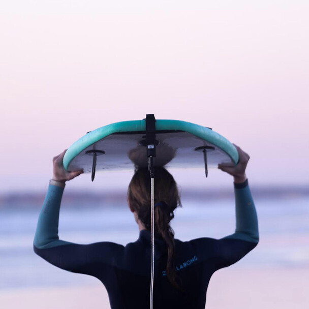 Surfen lernen Voraussetzungen, Surferin trägt Surfbrett auf dem Kopf mit Blick zum Meer