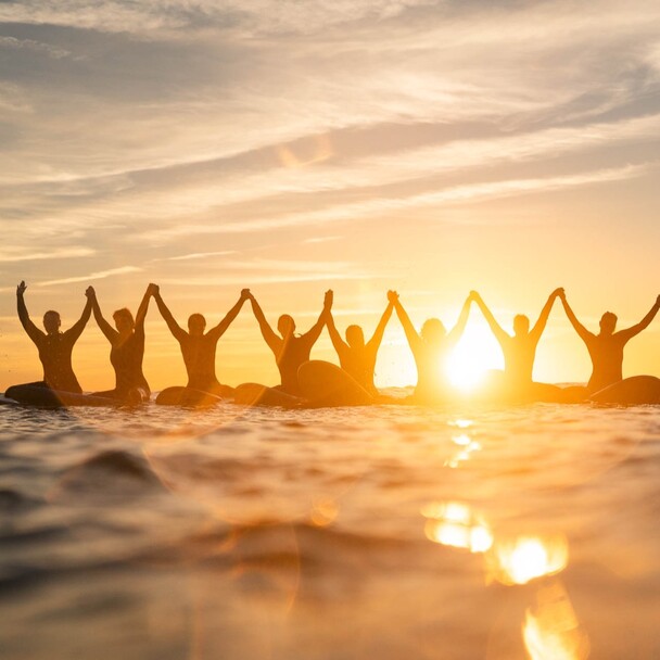 Learn to surf surf blog, group of surfers sitting in the water holding hands, sunset